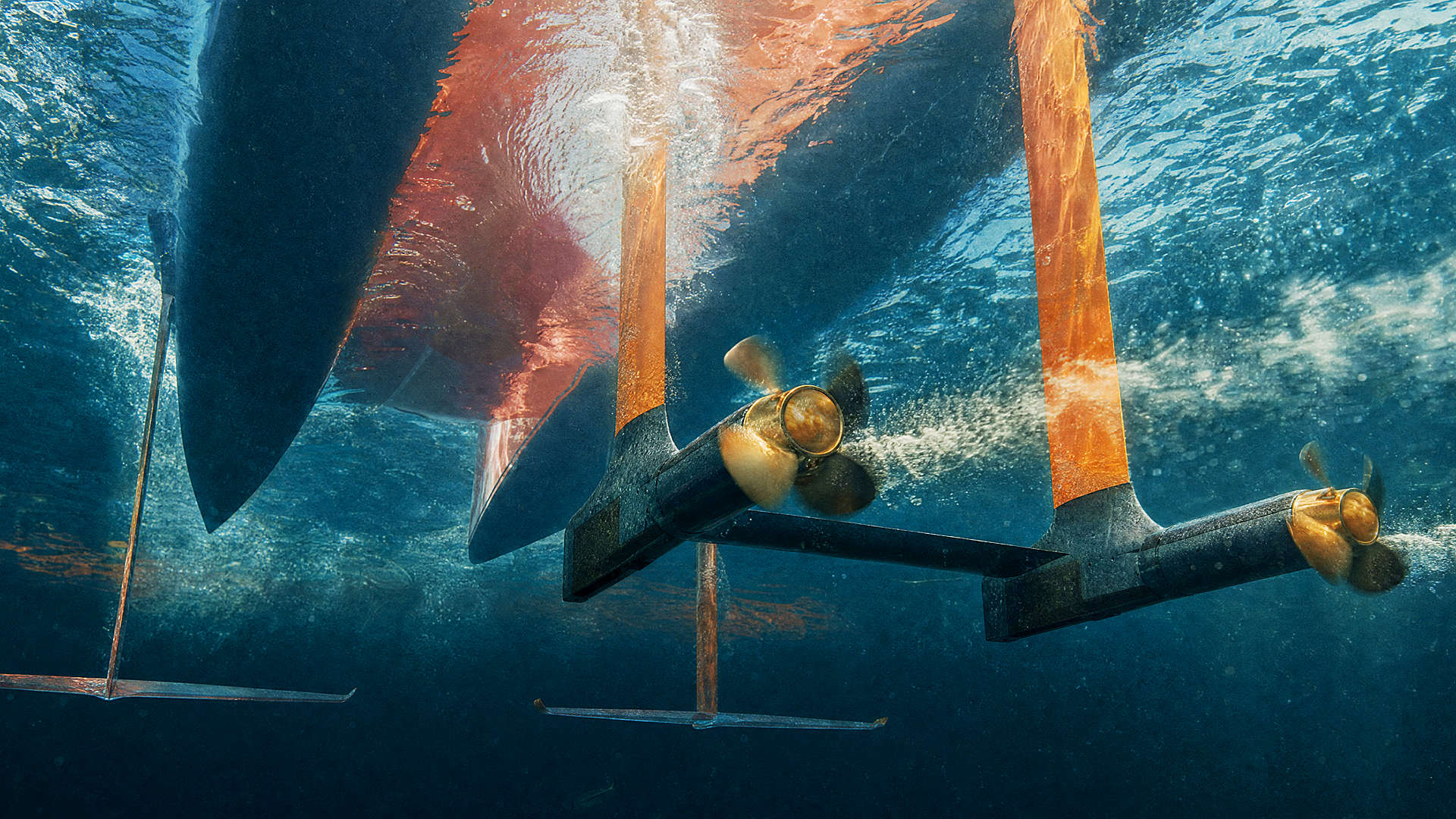 Underwater 3D render of the MobyFly MBFY-S powertrain, viewed from below the surface. Two retractable hydrofoil struts—rendered in blood orange—extend down from the hull, each terminating in a horizontal foil wing. Twin bronze propellers are mounted at the base of the struts, one in motion with visible cavitation. The hull rises above, partially visible through the refracted waterline. Deep blue water, dramatic upward lighting.